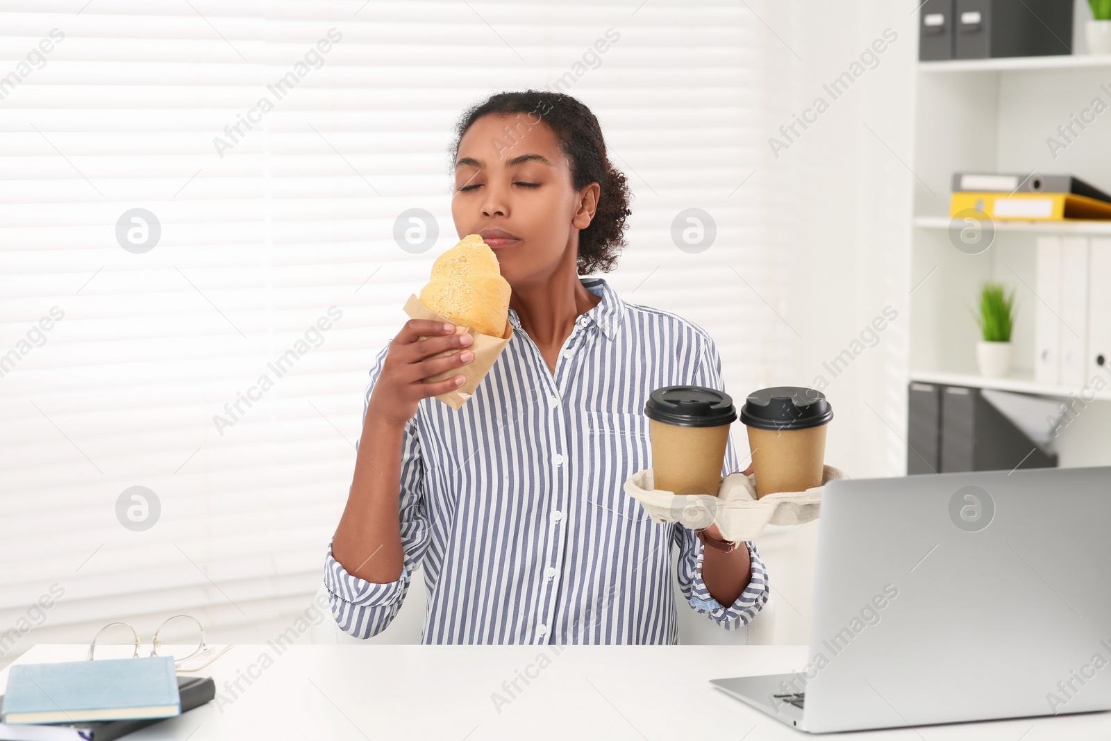Photo of African American intern with cups of drink eating croissant at white table in office