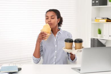 African American intern with cups of drink eating croissant at white table in office