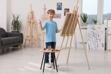Photo of Happy little boy showing thumbs up while painting in studio. Using easel to hold canvas