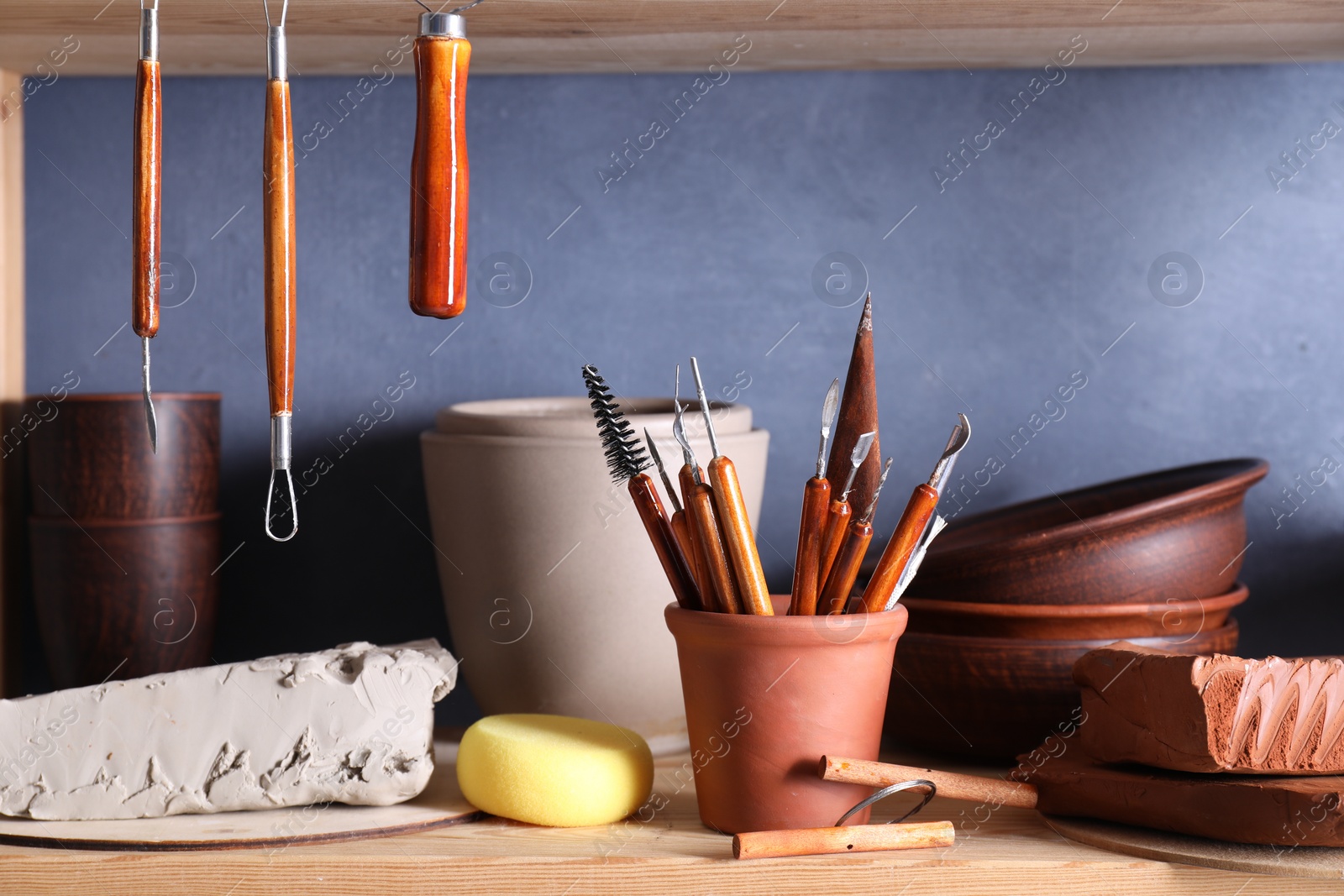 Photo of Set of different crafting tools and clay dishes on wooden rack in workshop, closeup