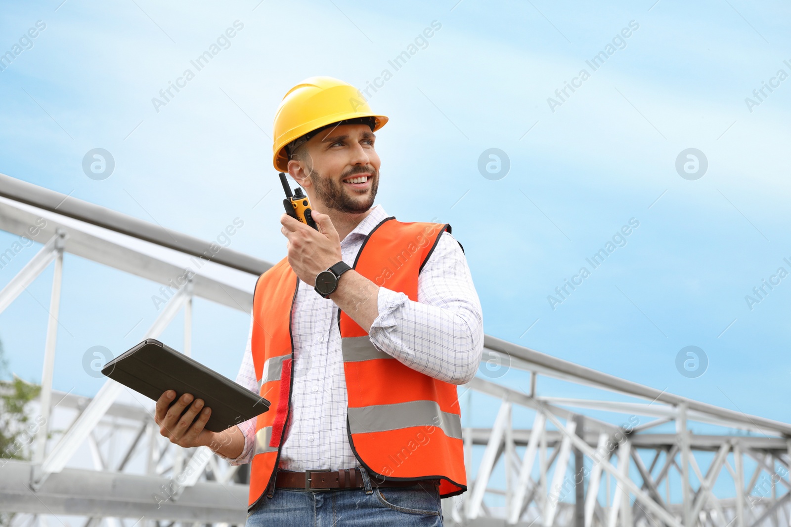 Photo of Professional engineer with tablet and walkie talkie near high voltage tower construction outdoors. Installation of electrical substation