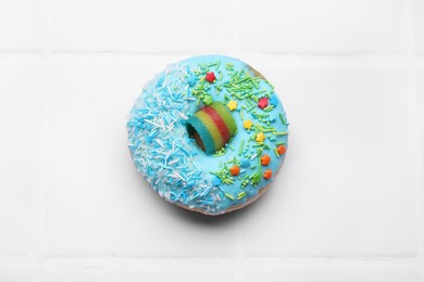 Photo of Glazed donut decorated with sprinkles on white tiled table, top view. Tasty confectionery