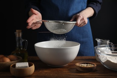 Photo of Making bread. Woman sifting flour over bowl at wooden table on dark background, closeup