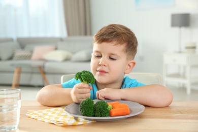 Adorable little boy eating vegetables at table in room