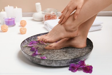 Photo of Woman soaking her feet in bowl with water and flowers on white wooden floor, closeup. Spa treatment