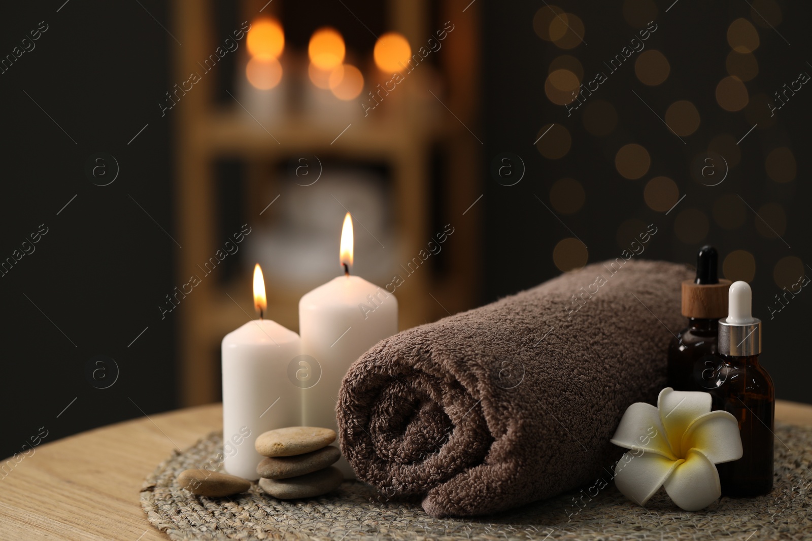 Photo of Spa composition. Rolled towel, cosmetic products, stones, burning candles and plumeria flower on table indoors, closeup. Space for text