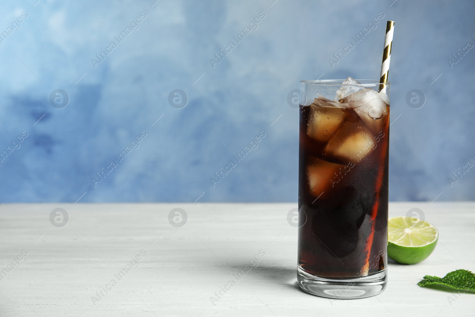 Photo of Refreshing soda drink with ice cubes and lime on white wooden table against blue background, space for text