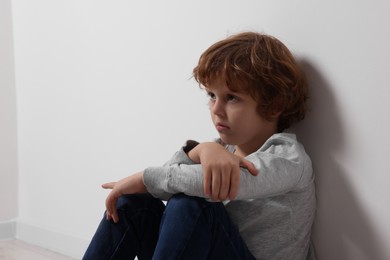 Photo of Child abuse. Upset boy sitting on floor near white wall