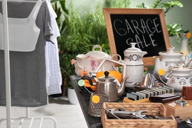 Photo of Blackboard with sign Garage Sale and many different stuff on black wooden table indoors