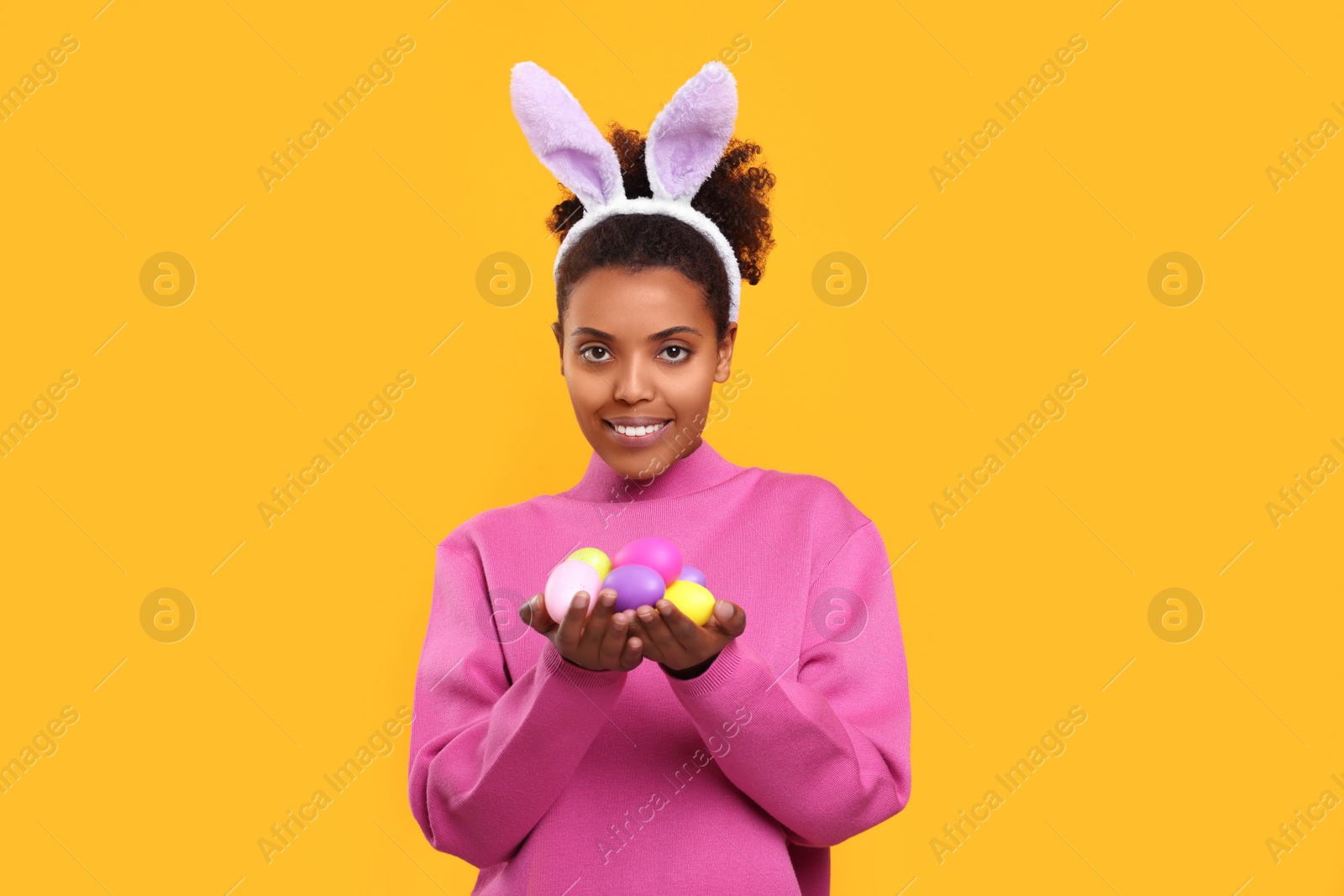 Photo of Happy African American woman in bunny ears headband holding Easter eggs on orange background