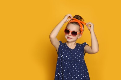 Photo of Cute little girl wearing stylish bandana and sunglasses on orange background, space for text