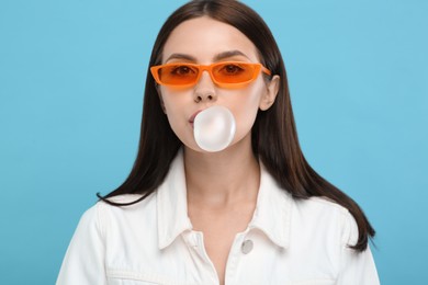 Beautiful woman in sunglasses blowing bubble gum on light blue background