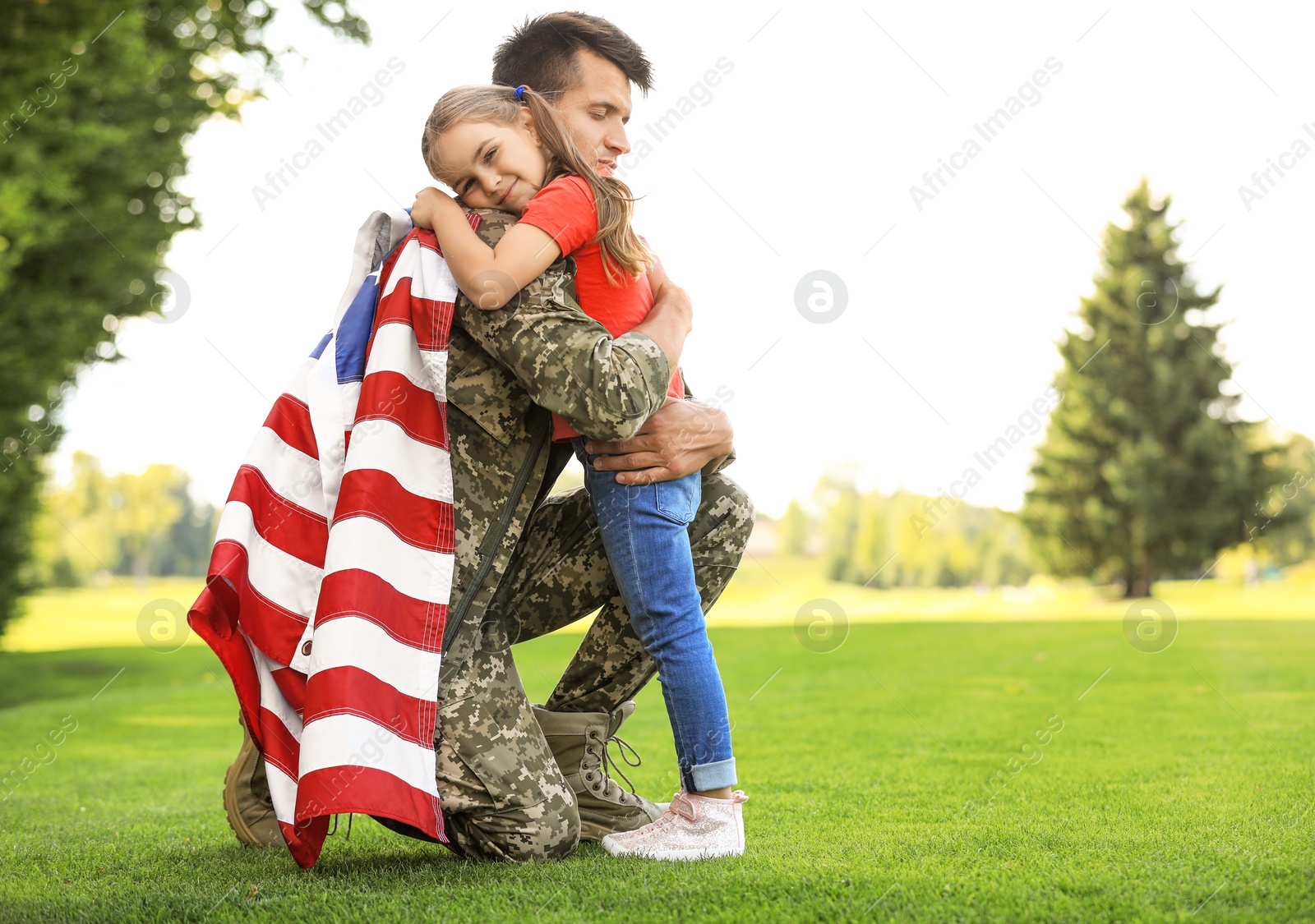 Photo of Father in military uniform with American flag hugging his daughter at sunny park