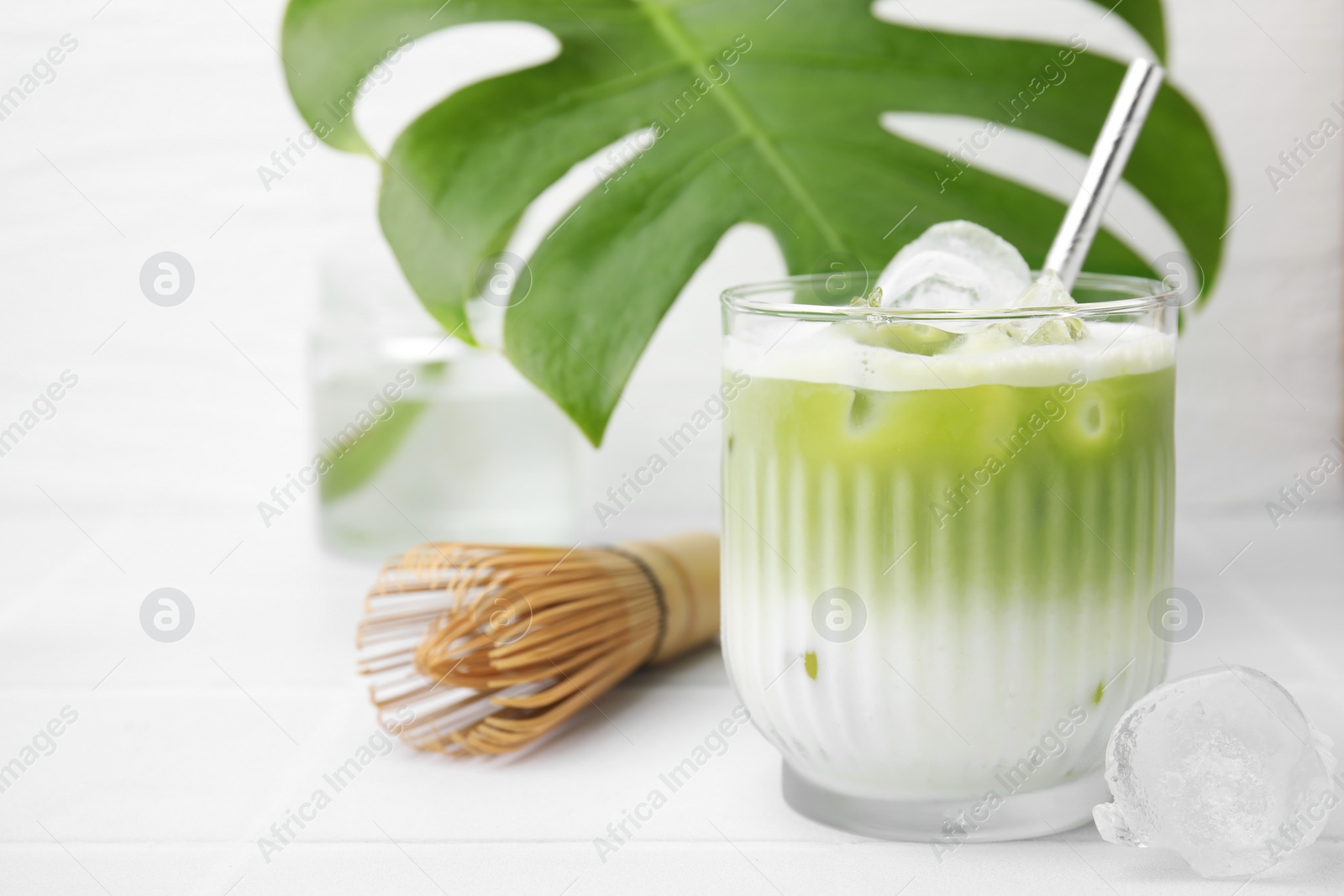 Photo of Glass of tasty iced matcha latte, bamboo whisk and leaf on white tiled table, closeup