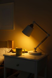 Photo of Stylish modern desk lamp, books and cup of drink on white nightstand in dark bedroom