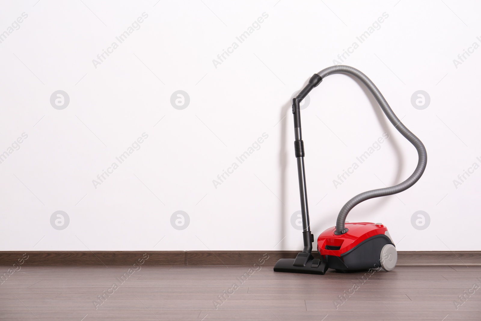 Photo of Modern red vacuum cleaner on wooden floor near white wall, space for text