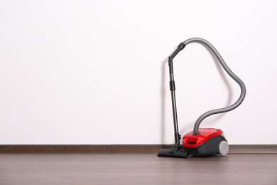 Modern red vacuum cleaner on wooden floor near white wall, space for text