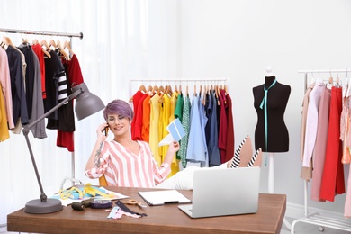 Photo of Beautiful young stylist talking on phone at workplace near rack with clothes