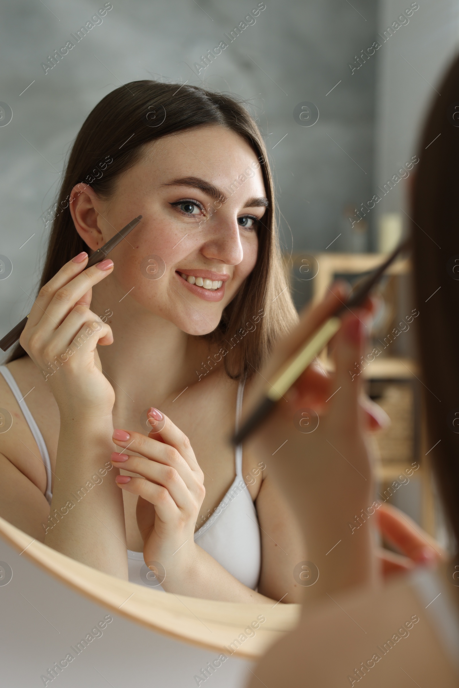 Photo of Smiling woman drawing freckles with pen near mirror indoors