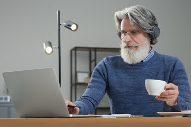 Photo of Middle aged man with laptop, headphones and cupdrink learning at table indoors