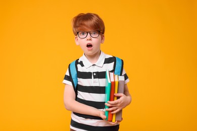Photo of Surprised schoolboy with backpack and books on orange background