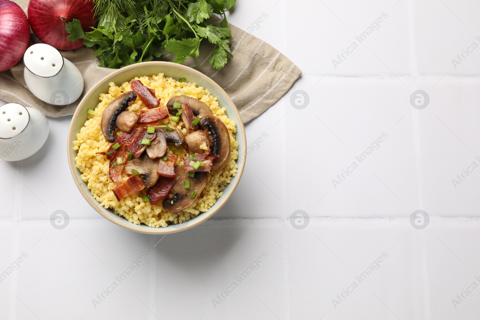 Photo of Tasty millet porridge with mushrooms, bacon and green onion in bowl on white tiled table, flat lay. Space for text