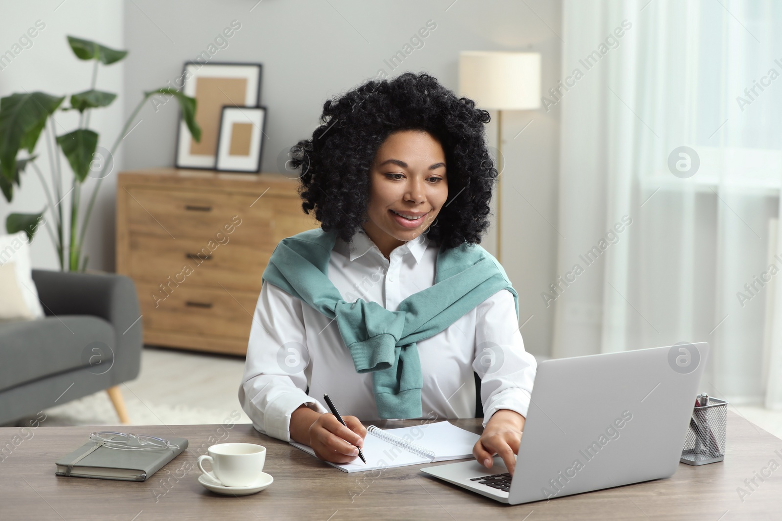 Photo of Happy young woman writing notes while using laptop at wooden desk indoors