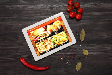 Modern tablet with open page for online food ordering surrounded by scattered products on wooden table, flat lay. Delivery service