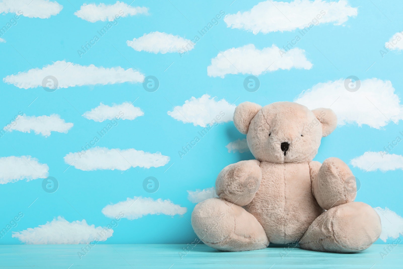 Photo of Teddy bear on wooden table near wall with painted blue sky, space for text. Baby room interior