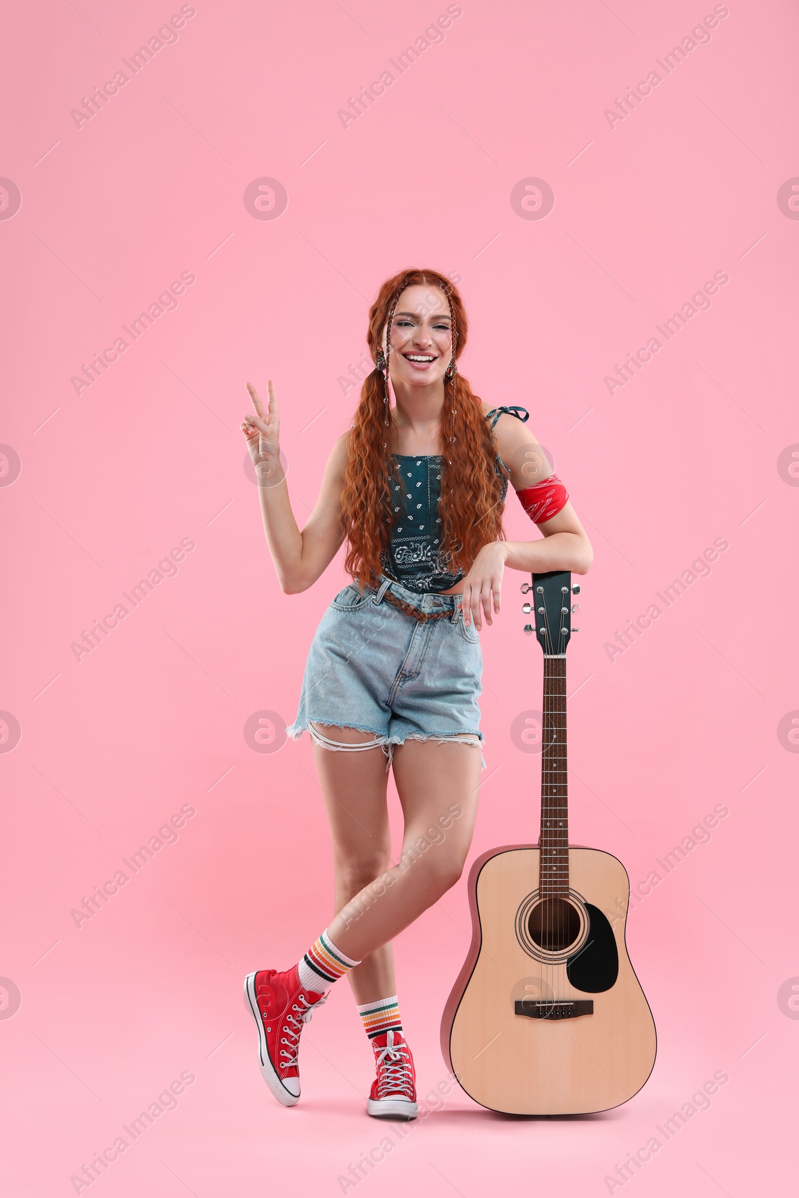 Photo of Stylish young hippie woman with guitar showing V-sign on pink background