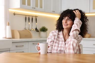 Beautiful young woman in stylish pyjama with cup of drink at wooden table in kitchen