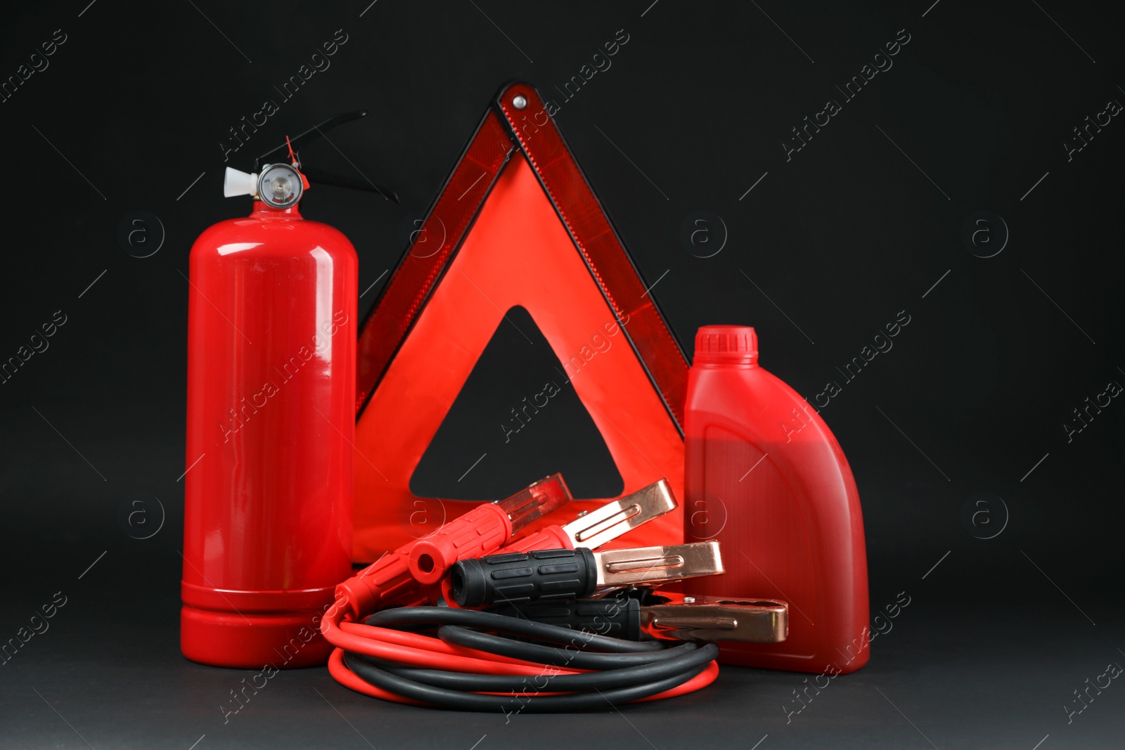 Photo of Emergency warning triangle, red fire extinguisher, battery jumper cables and motor oil on black background. Car safety