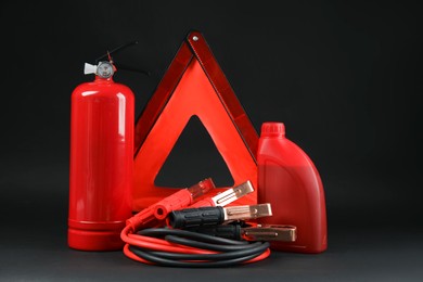 Photo of Emergency warning triangle, red fire extinguisher, battery jumper cables and motor oil on black background. Car safety