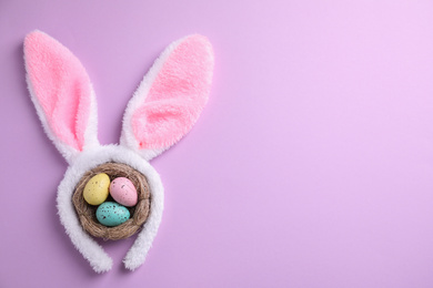 Photo of Headband with Easter bunny ears and dyed eggs in nest on violet background, flat lay. Space for text