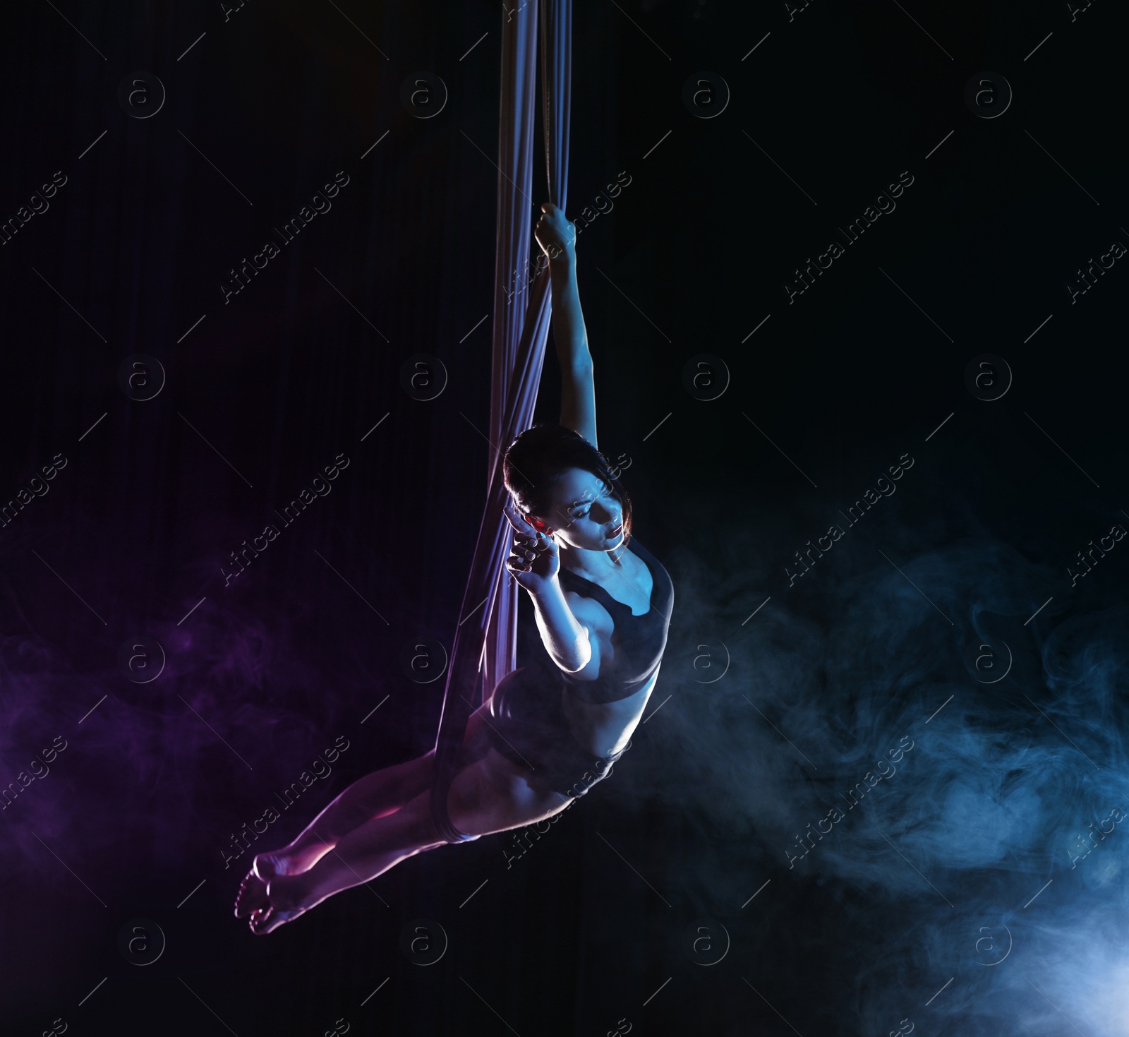 Photo of Young woman performing acrobatic element on aerial silk against dark background. Space for text