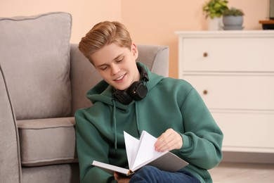 Photo of Teenage boy reading book near armchair in room