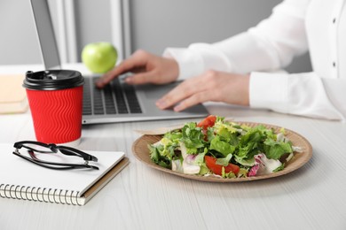 Photo of Office employee working with laptop at white wooden table, focus on vegetable salad and paper cup of coffee. Business lunch