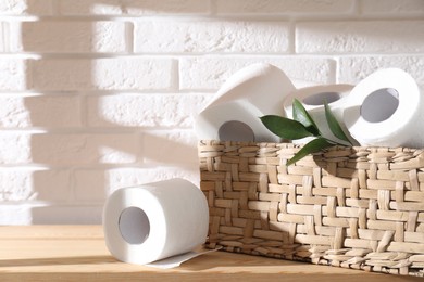 Photo of Toilet paper rolls in wicker basket and green leaves on wooden shelf against white brick wall