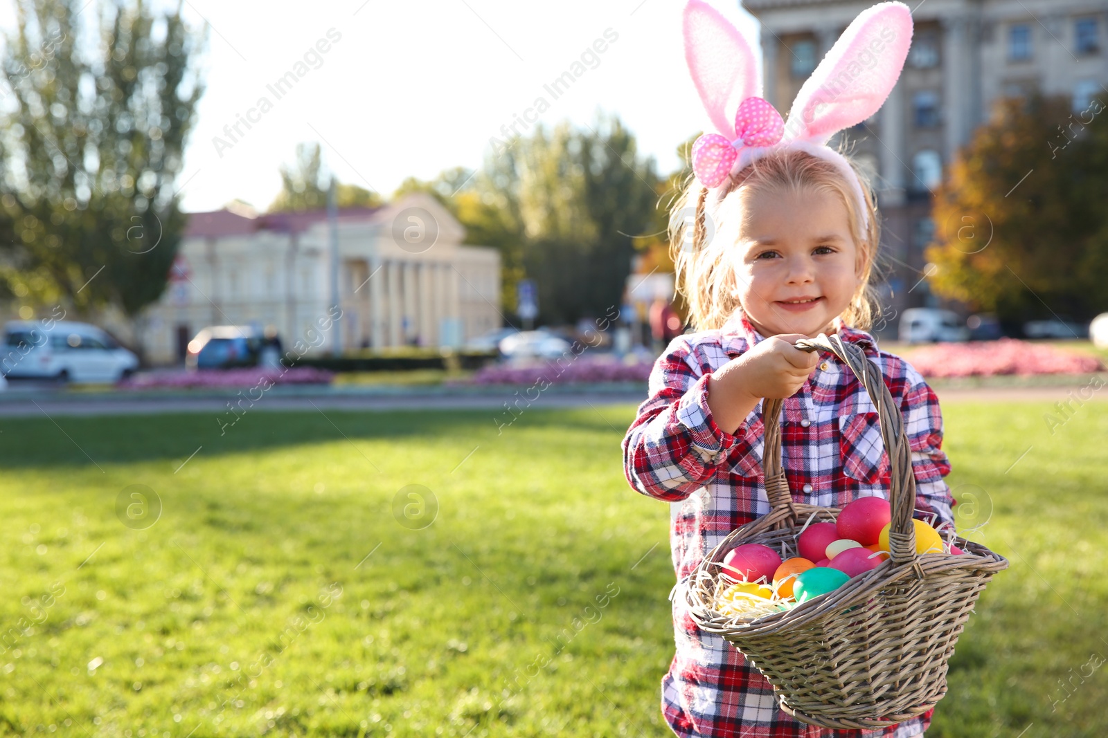 Photo of Cute little girl with bunny ears and basket of Easter eggs in park. Space for text