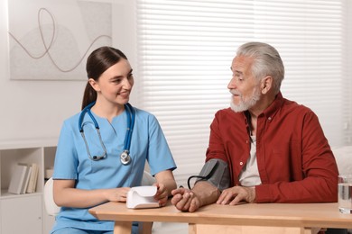Young healthcare worker measuring senior man's blood pressure at wooden table indoors