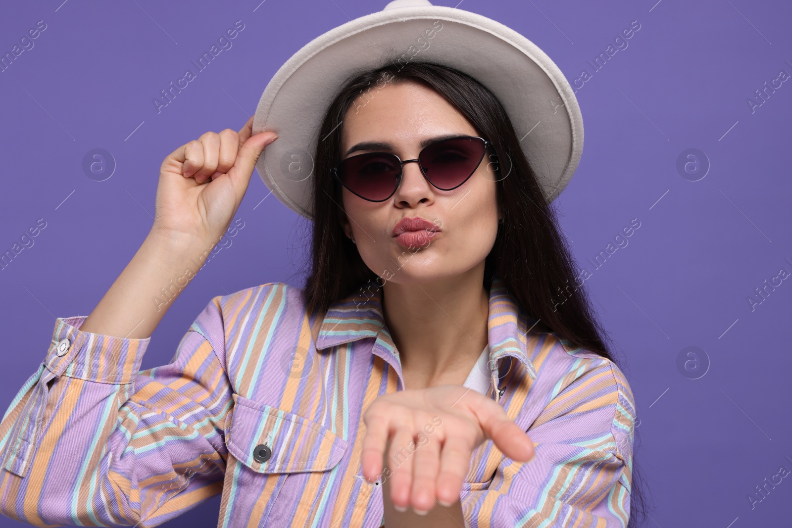 Photo of Beautiful young woman with stylish hat blowing kiss on purple background