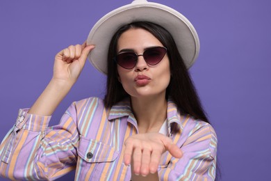 Beautiful young woman with stylish hat blowing kiss on purple background