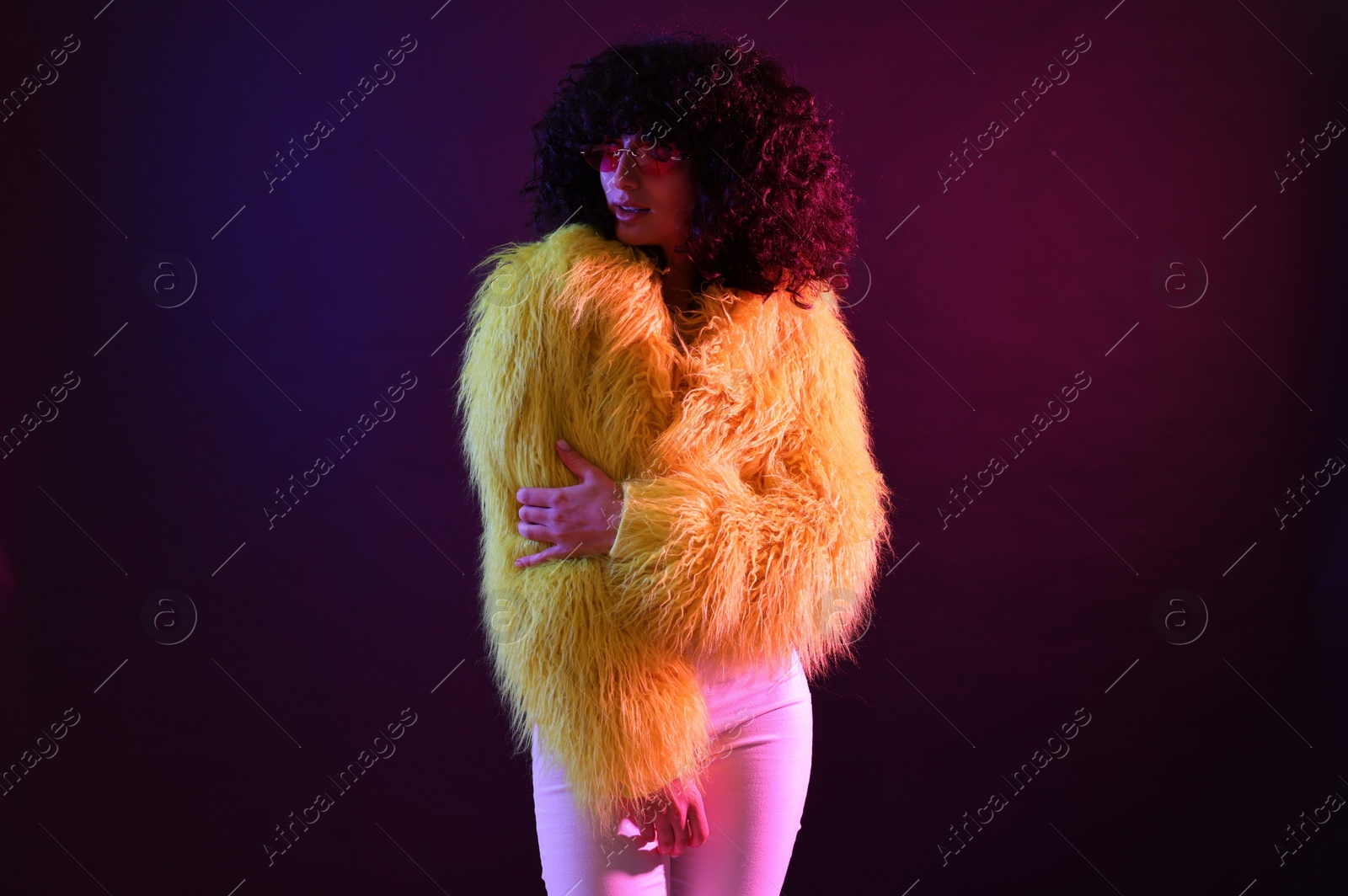 Photo of Beautiful young woman in yellow fur coat and sunglasses on color background in neon lights