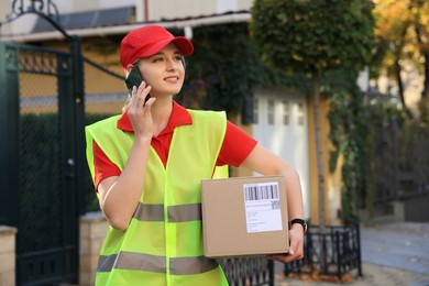 Photo of Courier in uniform with parcel talking on smartphone near private house outdoors