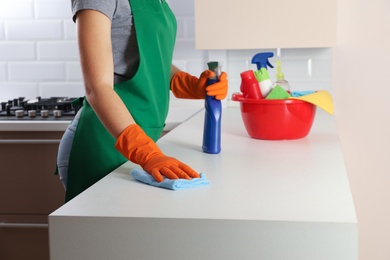 Photo of Woman in protective gloves cleaning kitchen table with rag, indoors