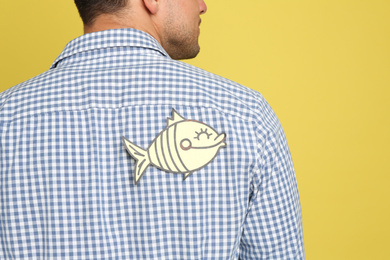 Photo of Man with paper fish on back against yellow background, closeup. April fool's day