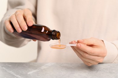 Woman pouring syrup from bottle into dosing spoon at table, closeup. Cold medicine