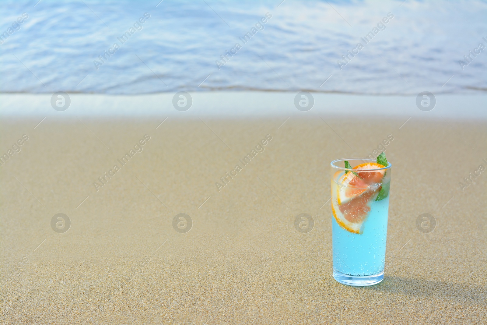Photo of Glass of refreshing drink with grapefruit and mint on sandy beach near sea, space for text