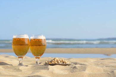 Glasses of cold beer and pistachios on sandy beach near sea, space for text