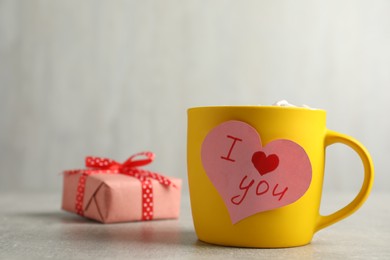 Heart shaped greeting card with phrase I Love You attached to yellow cup near gift box on table